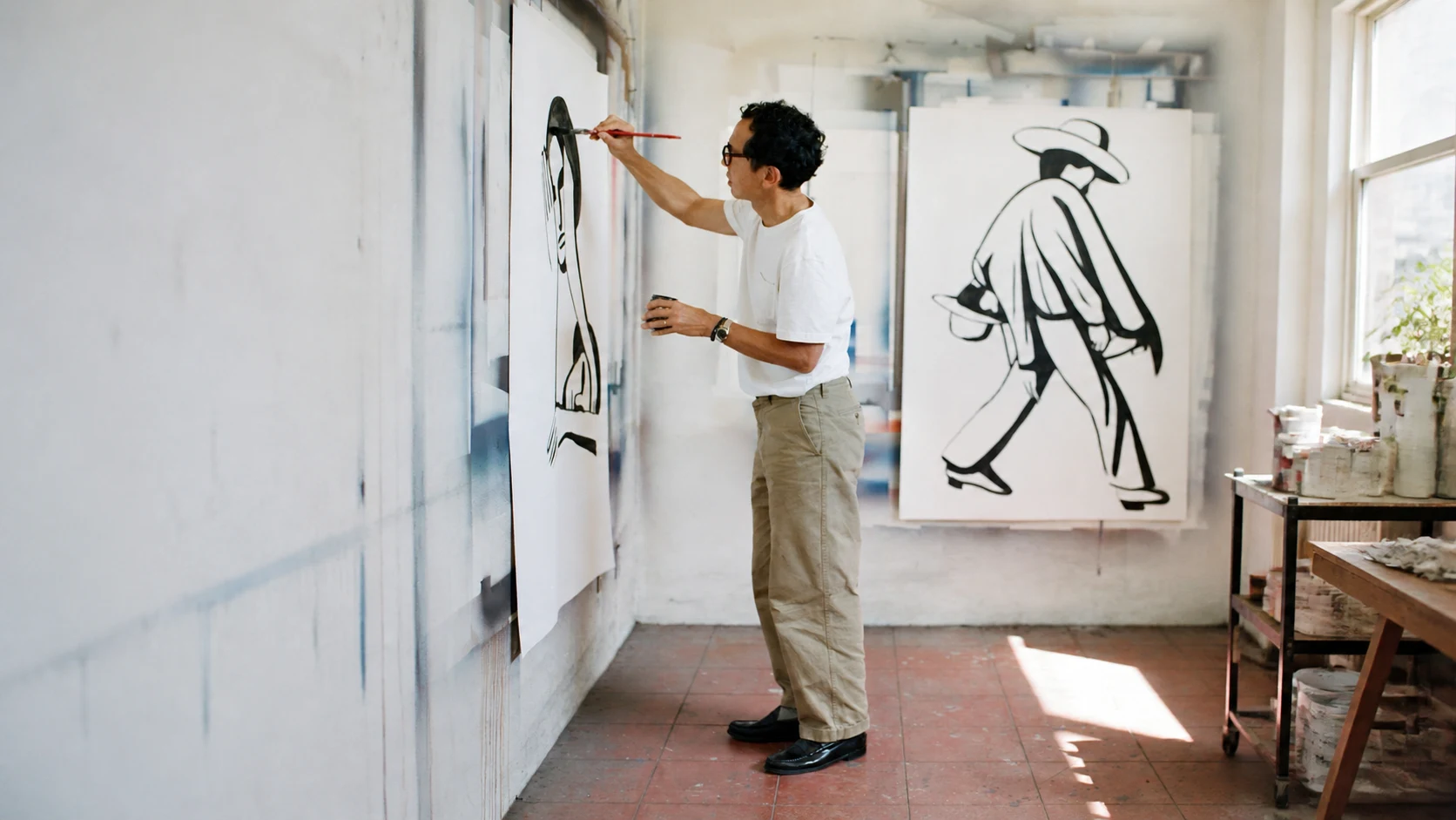 Elliot painting a large canvas, his full figure surrounded by artwork and materials in a sunlit studio.